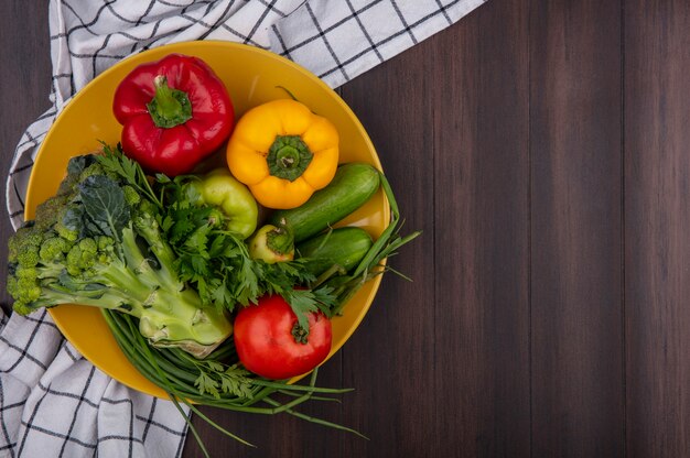 Top view  copy space colored bell peppers with broccoli  cucumber  tomato and green onion on yellow plate  on wooden background