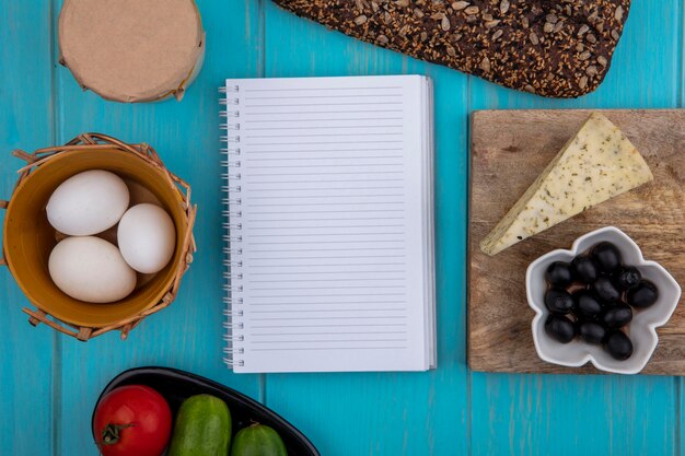 Top view  copy space copybook with black bread  cheese  olives  chicken eggs  yogurt and cucumbers with tomato on a turquoise background