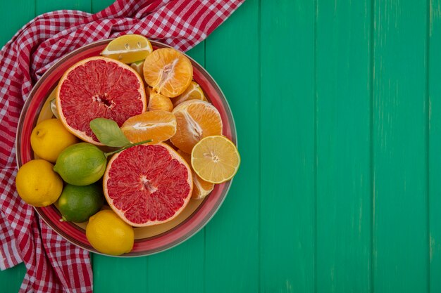 Top view  copy space cut in half grapefruit with peeled oranges and lemon with lime on a plate on a red checkered towel  on a green background