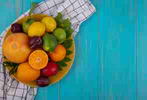 Free photo top view  copy space grapefruit with oranges  plums  lemons and limes on a yellow plate  on a checkered towel  on a turquoise background