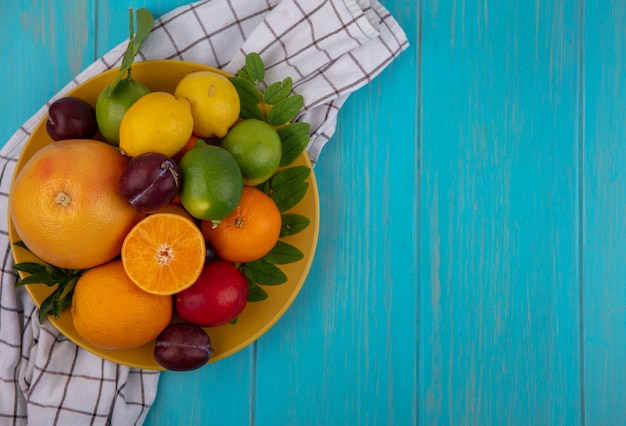 Top view  copy space grapefruit with oranges  plums  lemons and limes on a yellow plate  on a checkered towel  on a turquoise background