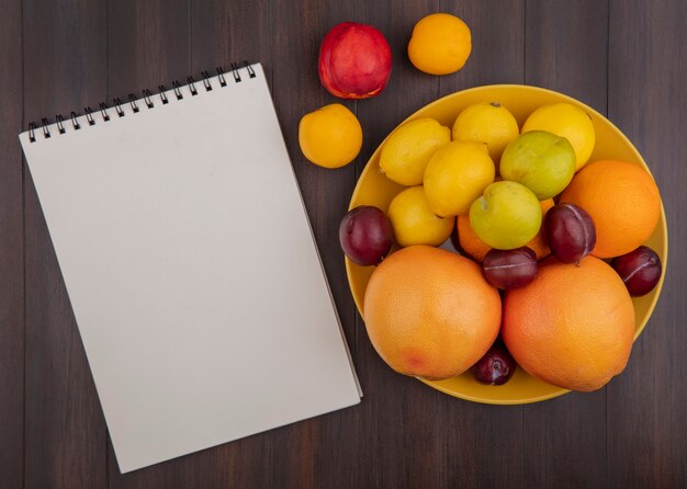 Top view  copy space lemons with oranges  plums and grapefruit in a yellow bowl with apricots and a peach with a notepad on a wooden background