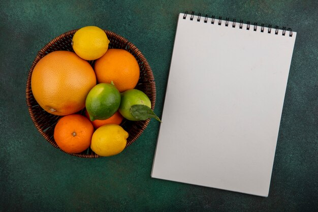 Top view  copy space oranges with grapefruit  lemons and limes in basket with notepad on green background