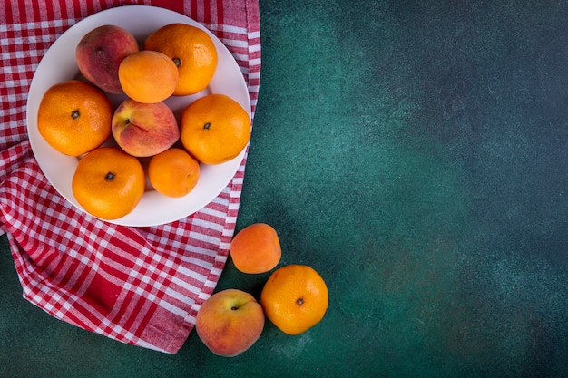 Free photo top view copy space peaches with tangerines and apricots on plate on kitchen towel on green