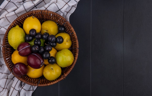 Top view  copy space plum with cherry plum and lemons with lime in a basket on a checkered towel  on a black background