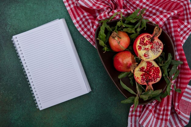 Top view  copy space pomegranate with twigs in a bowl with a checkered red towel and notepad on a green background