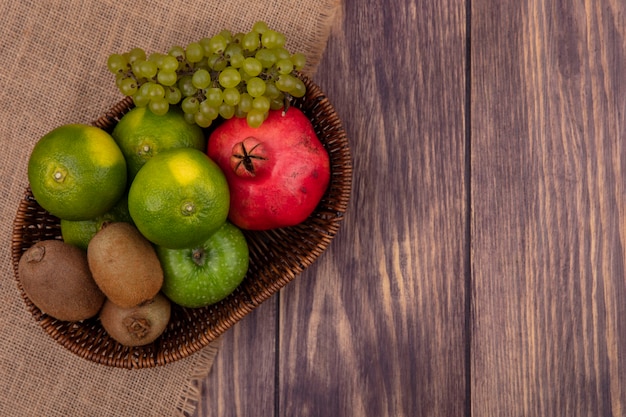 Top view copy space tangerines with grapes kiwi and pomegranate in a basket on a beige napkin on a wooden wall