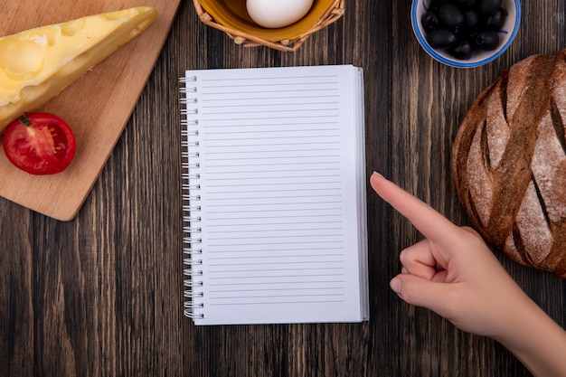 Top view  copy space  woman points to copybook with black olives with tomato  black bread and cheese on wooden background