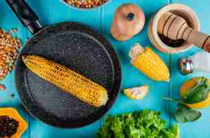 Free photo top view of corn cob in pan with corn seeds black pepper seeds cut corn salt lemon and lettuce around on blue surface