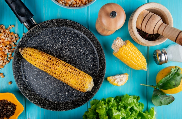 Top view of corn cob in pan with corn seeds black pepper seeds cut corn salt lemon and lettuce around on blue surface