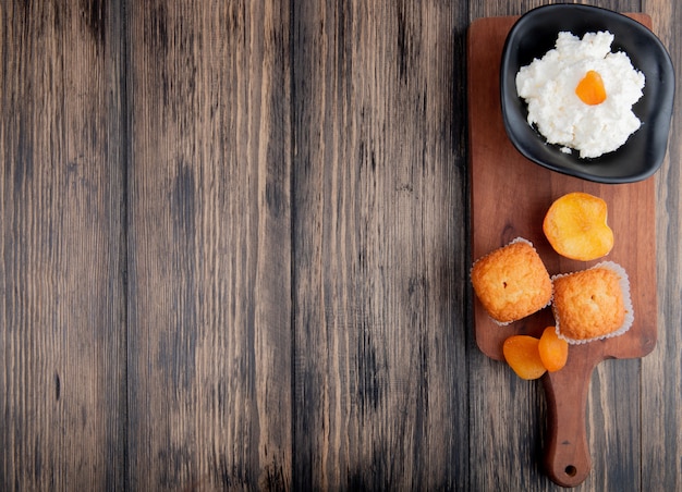 Top view of cottage cheese in a black bowl with muffins and dried apricots on wood cutting board on rustic with copy space