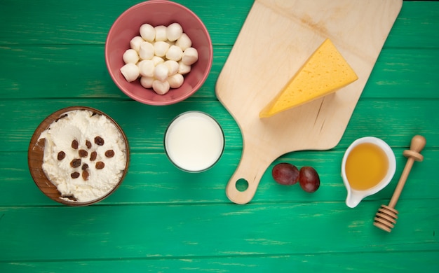 Top view of cottage cheese in a bowl topped with raisins  mozzarella and a piece of dutch cheese on wood cutting board with honey on green wood