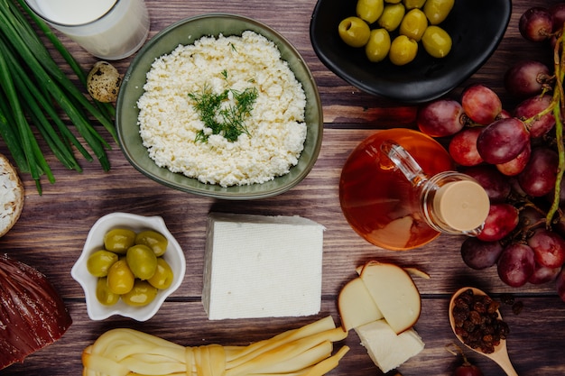 Top view of cottage cheese with herbs in a bowl and various kind of cheese with honey in a glass bottle  fresh grapes and pickled olives on rustic wood