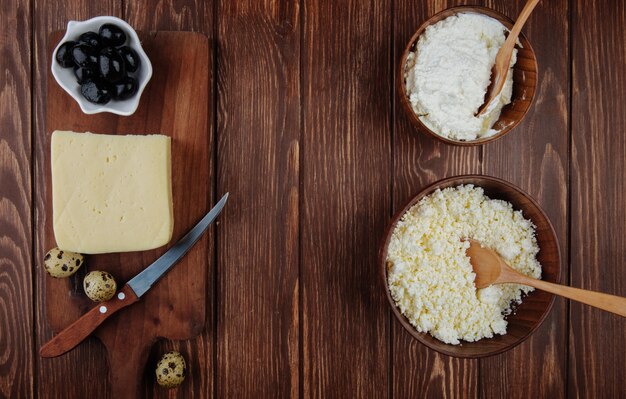 Top view of cottage cheese in wooden bowls and a piece of cheese on a wooden cutting board with a kitchen knife , quail eggs and pickled olives on rustic table