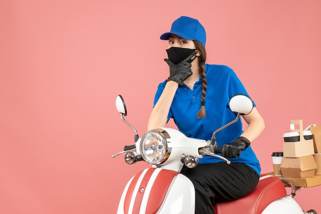 Вид сверху курьерской девушки в медицинской маске и перчатках, сидящей на скутере, доставляющей заказы на пастельном персиковом фоне