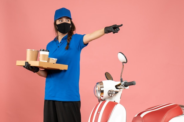 Top view of courier girl wearing medical mask gloves standing next to motorcycle holding coffee small cakes pointing forward on pastel peach color background