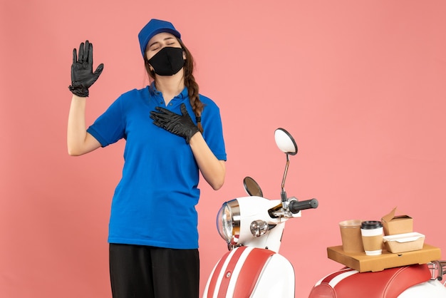 Top view of courier girl wearing medical mask gloves standing next to motorcycle with coffee cake on it dreaming on pastel peach color background