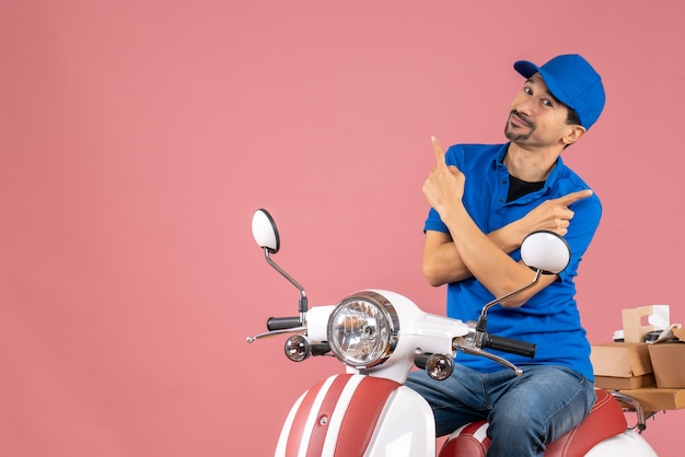 Top view of courier guy wearing hat sitting on scooter delivering orders pointing different sides on pastel peach background