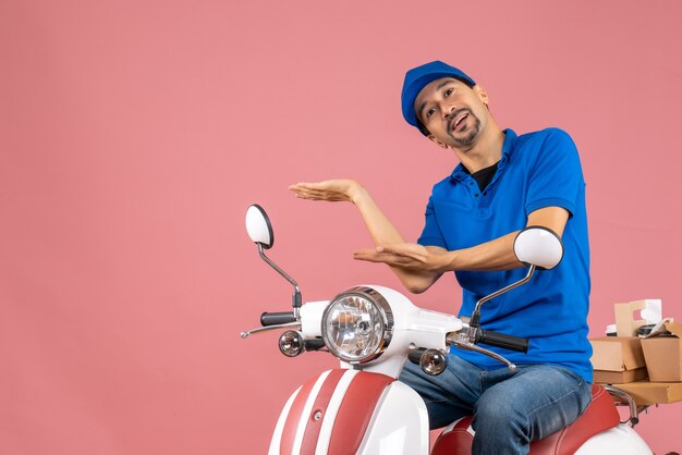 Top view of courier guy wearing hat sitting on scooter delivering orders pointing something on pastel peach background