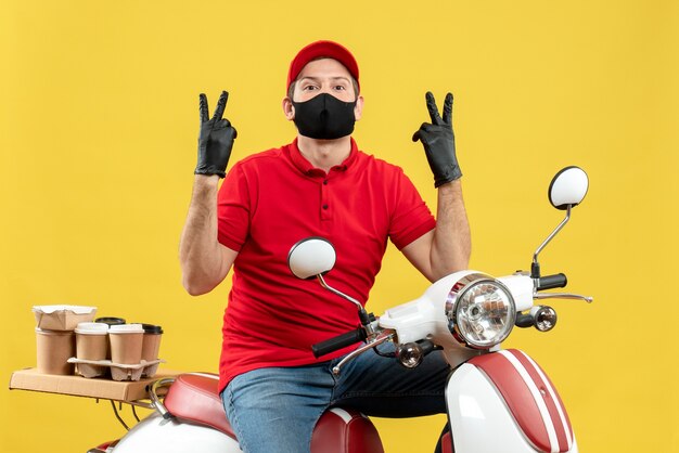 勝利のジェスチャーを作るスクーターに座って注文を配信する医療マスクで赤いブラウスと帽子の手袋を身に着けている宅配便の男の上面図