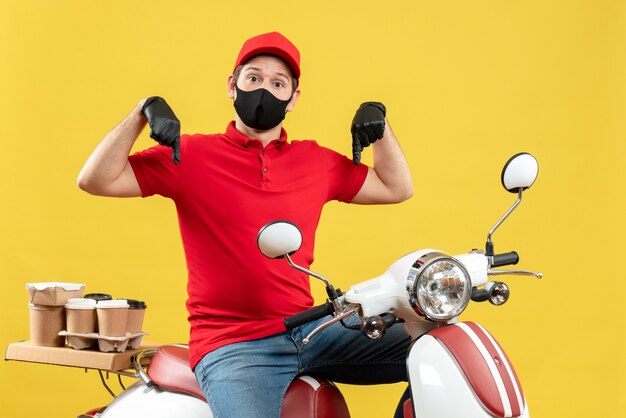 Top view of courier guy wearing red blouse and hat gloves in medical mask delivering order sitting on scooter pointing down