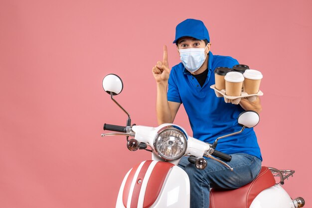 Top view of courier man in mask wearing hat sitting on scooter showing orders feeling satisfied on pastel peach background