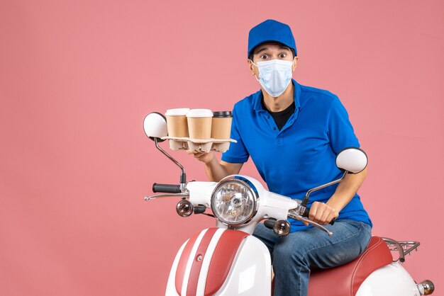 Top view of courier man in mask wearing hat sitting on scooter showing orders on pastel peach background