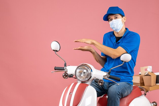 Top view of courier man in medical mask wearing hat sitting on scooter explaining something on pastel peach background