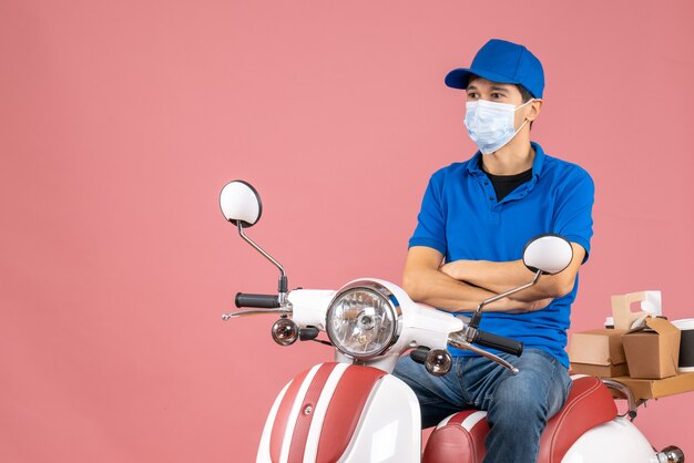 Вид сверху курьера в медицинской маске в шляпе, сидящего на скутере, внимательно смотрящего на что-то на пастельно-персиковом фоне