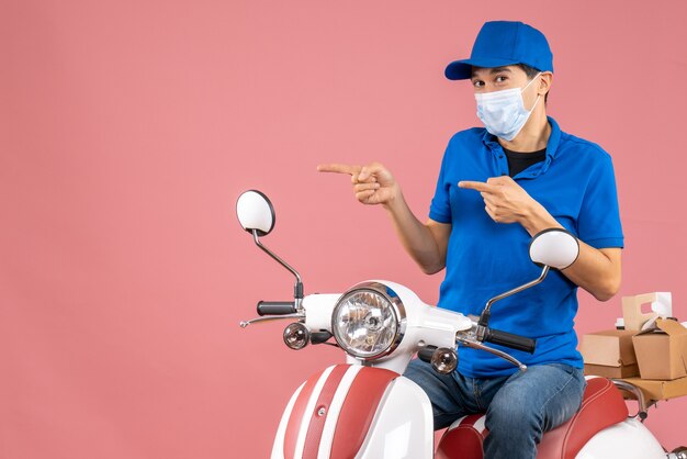 Top view of courier man in medical mask wearing hat sitting on scooter on pastel peach background