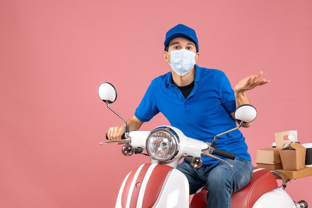 Top view of courier man in medical mask wearing hat sitting on scooter wondering something on pastel peach background