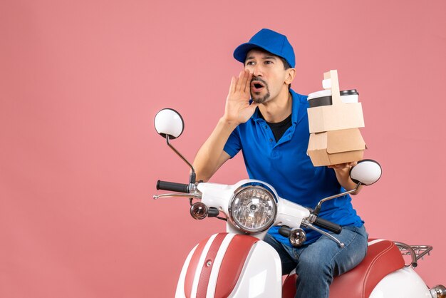 Top view of courier man wearing hat sitting on scooter holding orders calling someone on pastel peach background