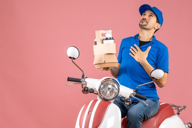 Top view of courier man wearing hat sitting on scooter holding orders dreaming on pastel peach background