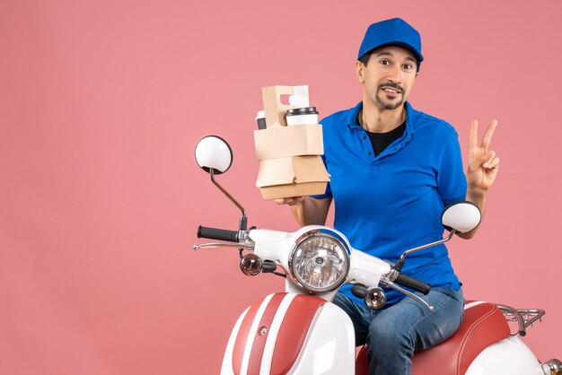 Top view of courier man wearing hat sitting on scooter showing orders making victory gesture on pastel peach background