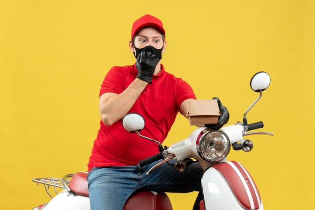Top view of courier man wearing red blouse and hat gloves in medical mask sitting on scooter holding order feeling surprised
