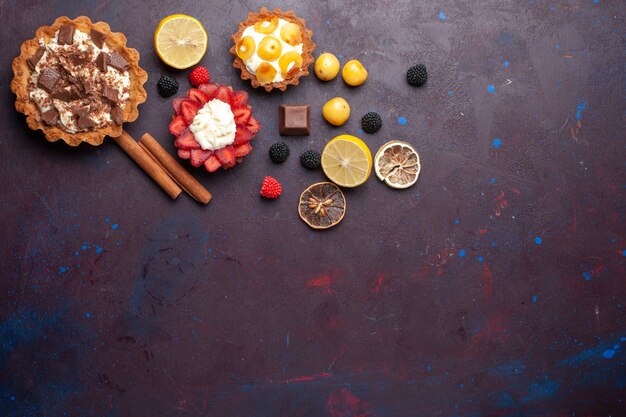 Top view of creamy cakes with fruits and berry confitures on dark-purple surface