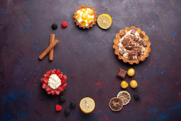 Top view of creamy cakes with fruits and berry confitures on the dark surface