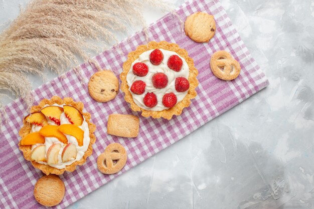 Вид сверху на сливочные пирожные с белым вкусным кремом и нарезанными клубникой, персиками, абрикосами с печеньем на светлом столе, выпечка с фруктовым кремом