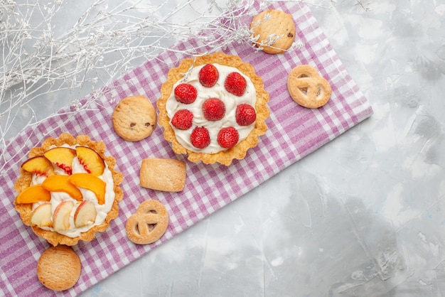 Вид сверху сливочных пирожных с белым вкусным кремом и нарезанной клубникой, персиками, абрикосами с печеньем на светлом столе, выпечка с фруктовым кремом