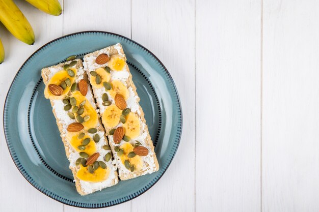 Top view of crispy crackers with cream cheese, slices of banana, almond and pumpkin seeds on a plate on white wood with copy space