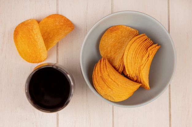 Top view of crispy potato chips on a bowl with a glass of cola on a beige wooden table