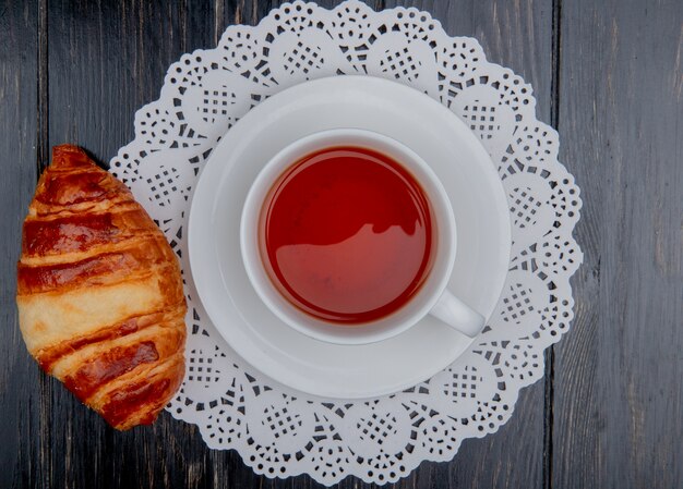 top view of croissant and cup of tea on teabag on paper doily and wooden background