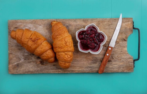 Top view of croissants and raspberry jam with knife on cutting board on blue background