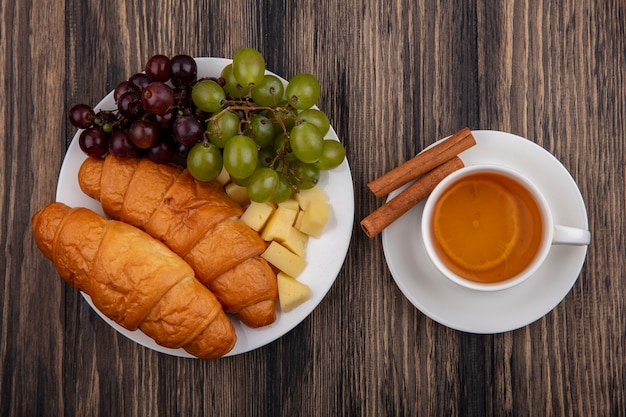 Top view of croissants with grapes and cheese slices in plate with cup of hot toddy with cinnamon on saucer on wooden background