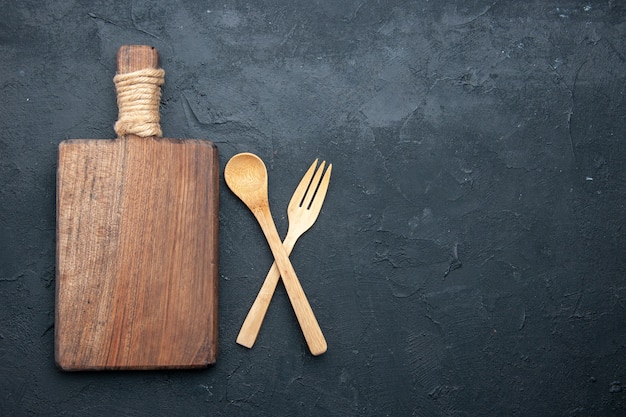 Top view crossed wooden spoon and fork serving board on dark table with copy place