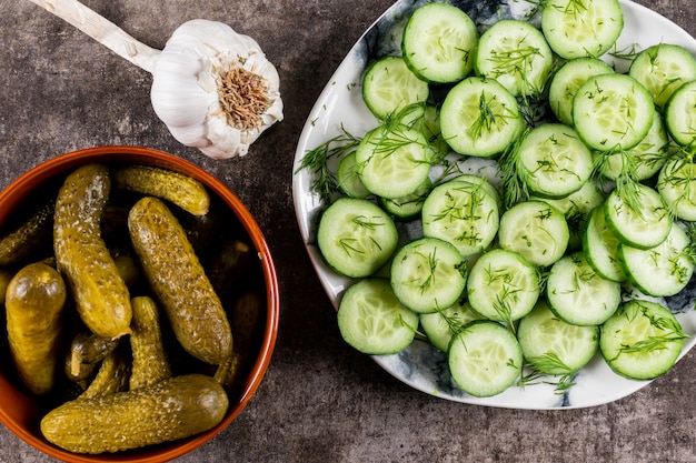 Top view cucumber fresh sliced with dill on plate and pickled in bowl on brown stone