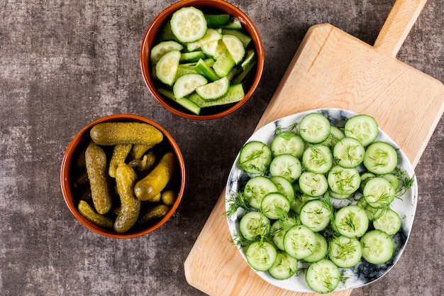 Top view cucumber fresh sliced with dill on plate on wooden cutting board and pickled in bowl on brown stone