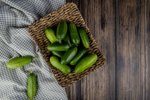 Top view of cucumbers in basket plate with other ones on cloth and wooden surface with copy space