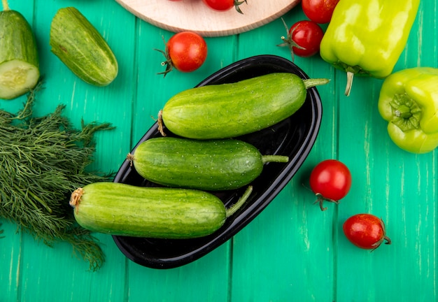 Top view of cucumbers in bowl and pepper tomato dill around on green