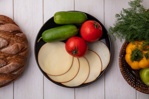 Вид сверху огурцы с болгарским перцем и укропом в корзине с копченым сыром и помидорами с буханкой хлеба на белом фоне